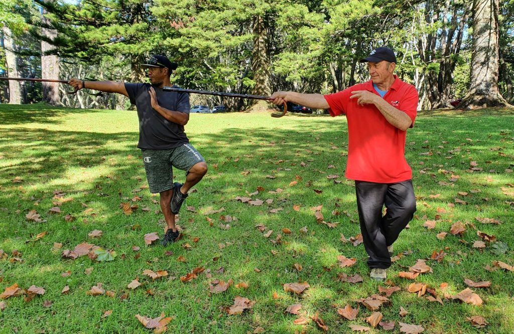 Two men, each balanced on one leg while pointing a wooden walking stick.