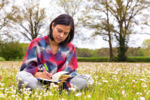 Woman writing in a diary, while sitting cross legged on the grass, with daisies growing around her