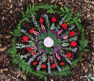 A symmetrical, circular design made of green and purple leaves, red flowers, and sprigs of rosemary, all enclosed in a circlet of leafy, green ferns.