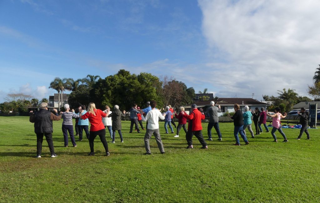 Group of people doing tai chi in a park, with blue skies overhead and a few white clouds overhead.
